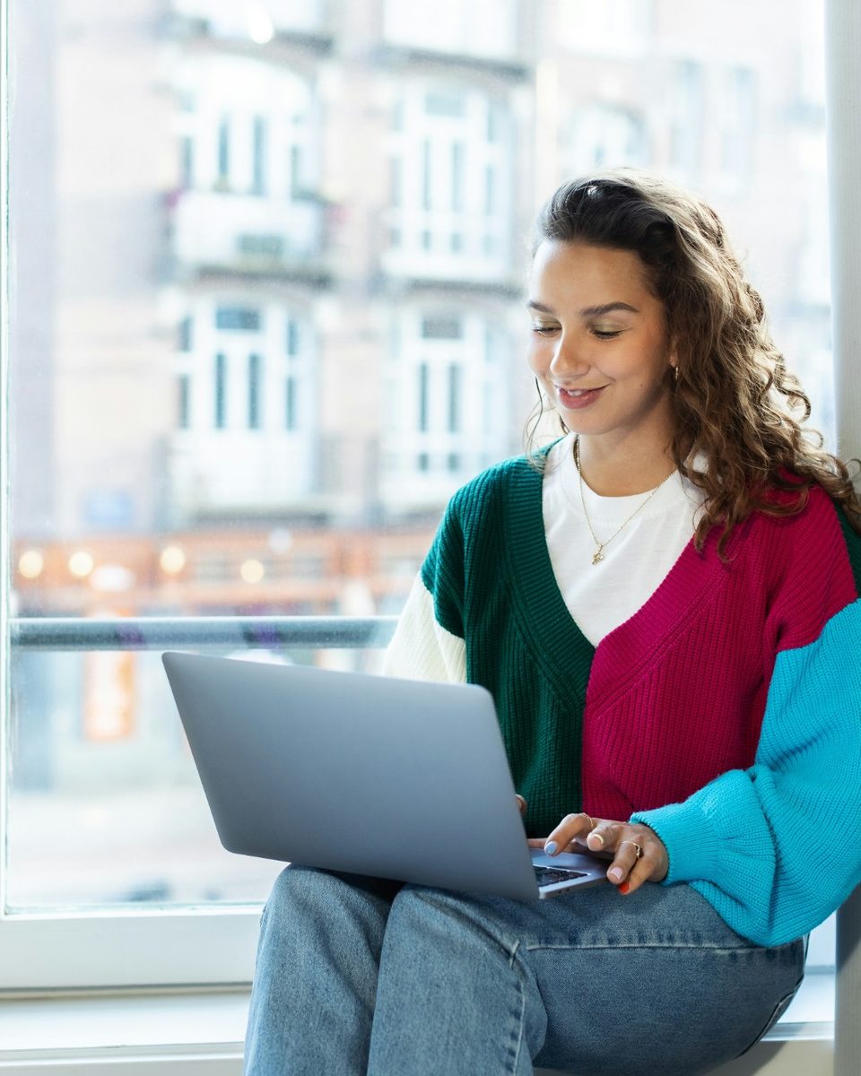 A woman sitting on a windowsill, working on her laptop