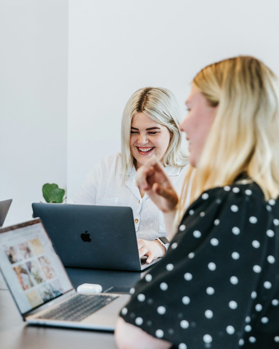 Zwei Frauen arbeiten lachend gemeinsam an einem Tisch mit Laptops