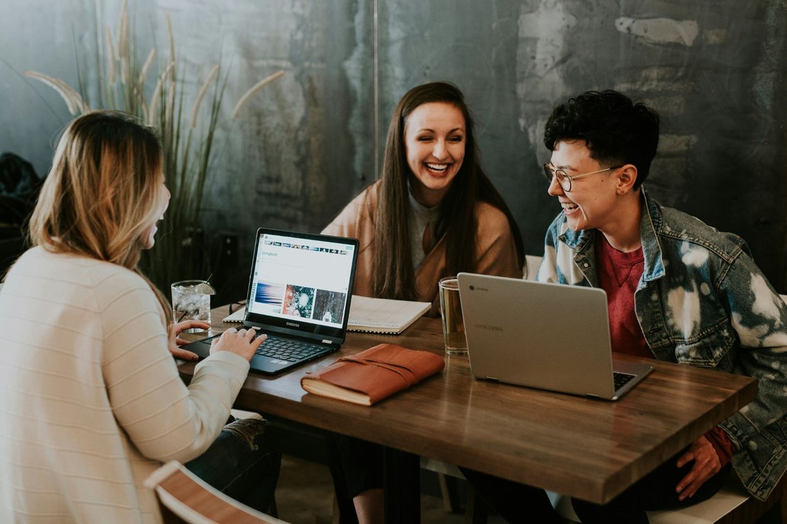 Drei Menschen sitzen lachend mit Laptops an einem Tisch